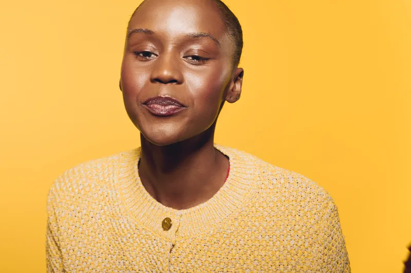 Portrait of a confident black woman with dark skin wearing a yellow knitted sweater, smiling subtly against a bright yellow background. Concept of beauty, fashion, and self-confidence.