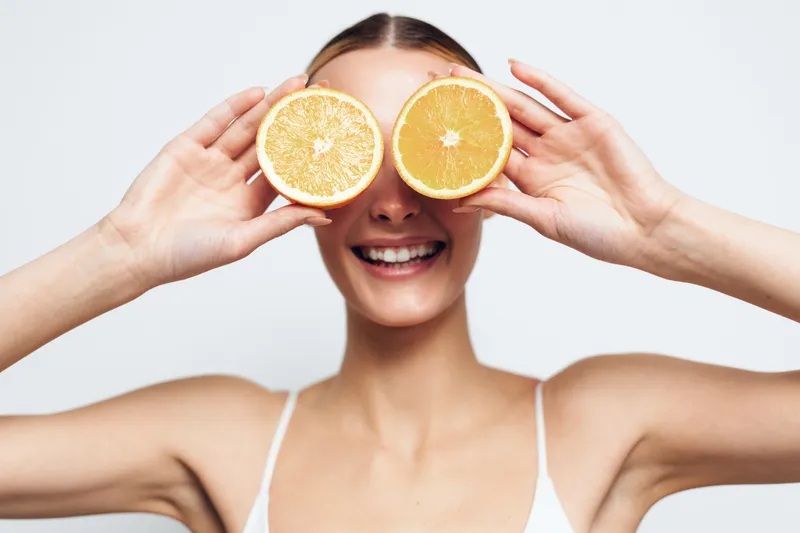 Happy smiling woman holds two halves of fresh orange fruit covering her eyes isolated on white background with positive expression and healthy skin closeup portrait.