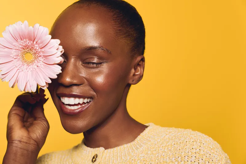 Happy African woman with dark skin holding pink flower near face smiles with closed eyes. Studio portrait on yellow background. Concept of beauty, skincare, wellness, and natural happiness.