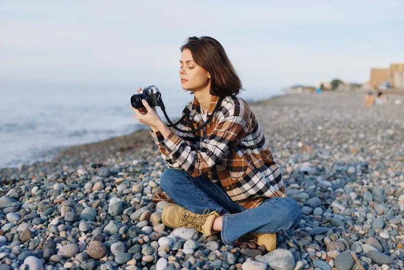 Photographer on a pebbled beach with camera, seated and focused, capturing outdoor scenery, calm moment, and natural light during a quiet day, horizon.