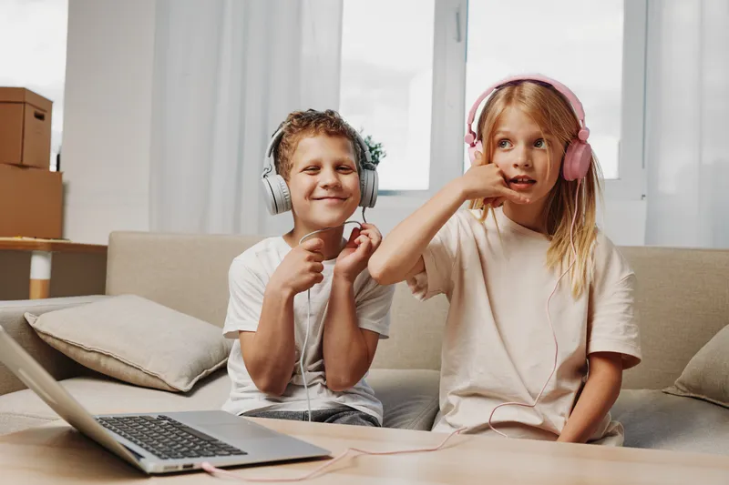 Two children with headphones enjoy music and play with gestures sitting on a couch near a laptop at home.