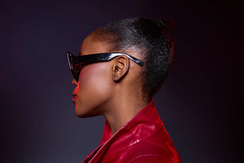 Profile of a young African woman with dark skin wearing black sunglasses and a red leather jacket against a dark background. Concept for fashion, style, and confidence.