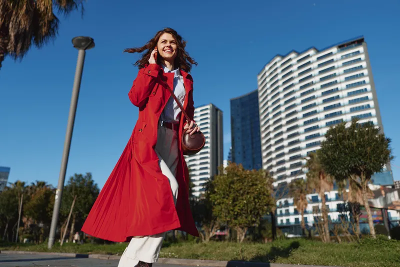 Young adult woman with curly hair in a red trench coat walks outdoors on a sunny city street, smiling as modern high-rise buildings rise behind her.