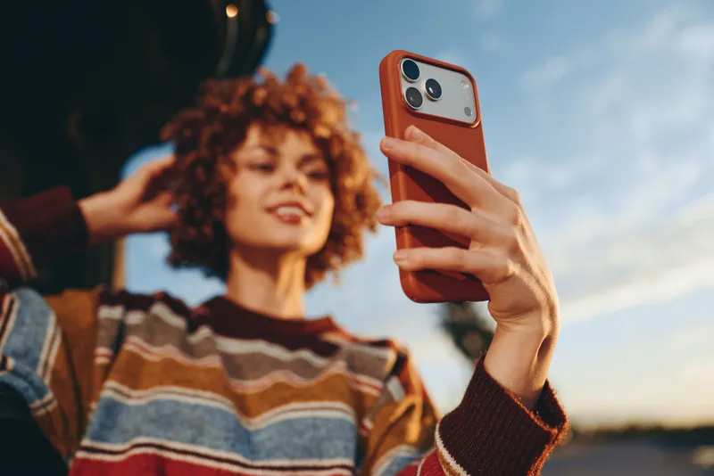 Woman lifestyle selfie using smartphone outdoors, hand holds red phone case while curly haired woman smiles in blurred background, casual sweater, warm sunset light, beach.