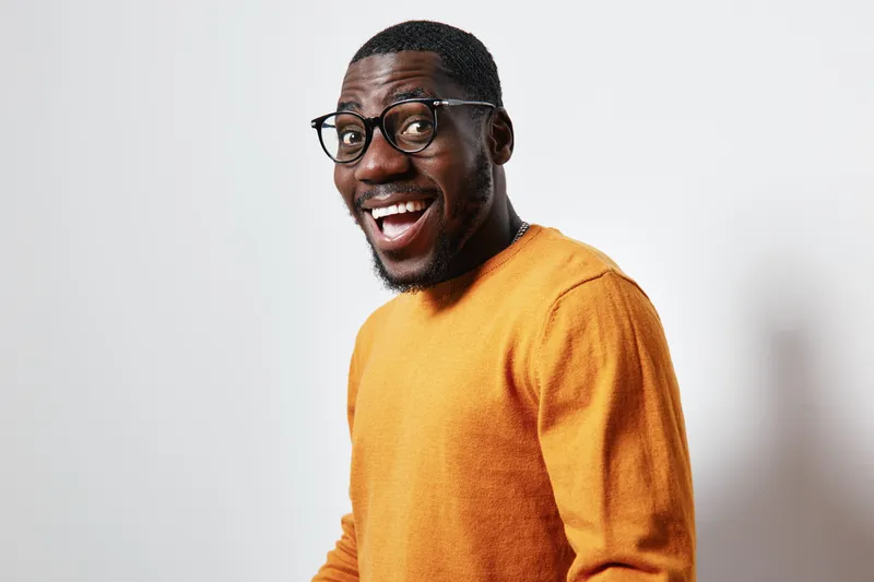 African American man with dark skin wearing glasses and orange sweater smiling happily on white background. Portrait of cheerful black male with beard and short hair in casual clothing.