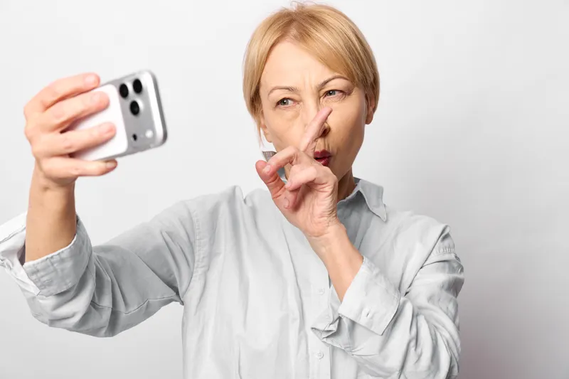 Surprised mature woman takes a selfie with smartphone, covers lips with finger in silence gesture, gray shirt on plain white background, indoor portrait, worried expression.