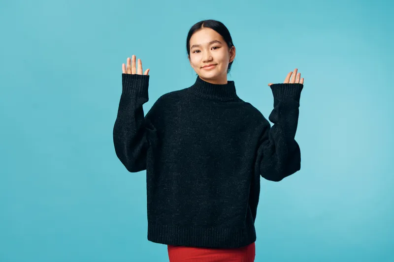 Young woman wearing black sweater standing with hands raised smiling at camera on blue background. Casual style, friendly gesture and confident expression in studio portrait.