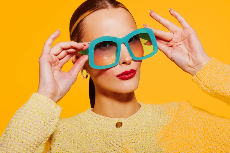 Stylish woman in yellow sweater adjusts large turquoise sunglasses on bright orange background. Fashion portrait with vibrant colors and confident expression. Concept of summer style and accessories.