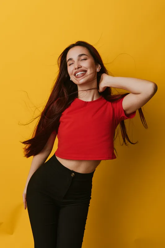 Young woman with long hair wearing red crop top and black pants smiling with eyes closed posing against yellow background in casual style with joyful expression and relaxed posture.