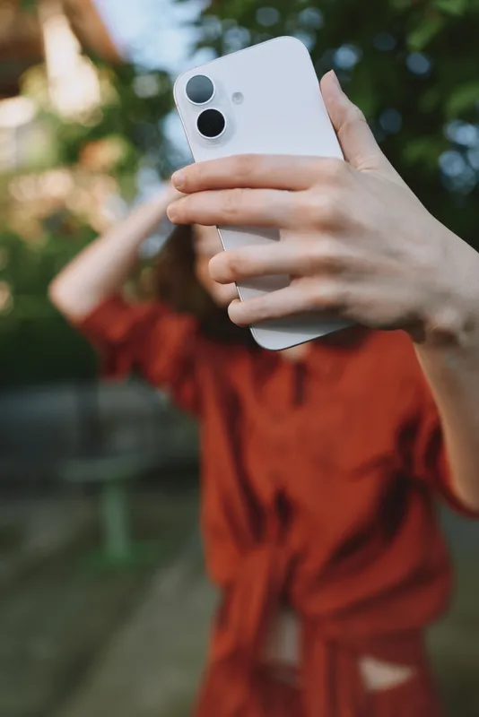 Young woman takes a candid outdoor selfie with a white smartphone, standing in a sunlit garden and enjoying a relaxed, casual moment with a friendly vibe