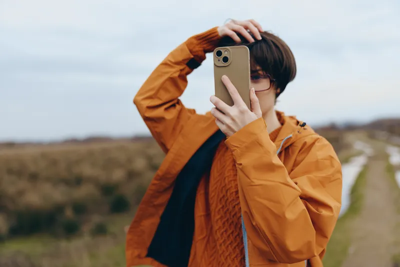 Woman in orange jacket and glasses outdoors holding a smartphone for a selfie on a country road. Lifestyle travel vibe, natural light, blurred countryside background, relaxed pose today.