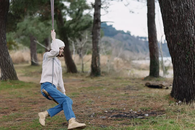 Swinging through a pine forest, a person enjoys outdoor recreation and relaxation on a simple swing among trees, grass, and tranquil nature, capturing a candid moment of leisure and carefree motion.