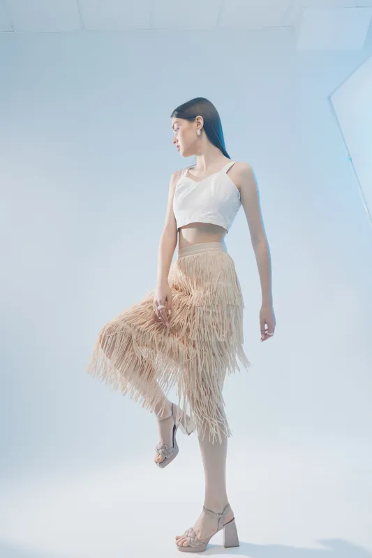 Young woman in stylish beige fringe skirt and white crop top poses gracefully against a light background. Concept of woman beauty fashion with elegance and modern style.