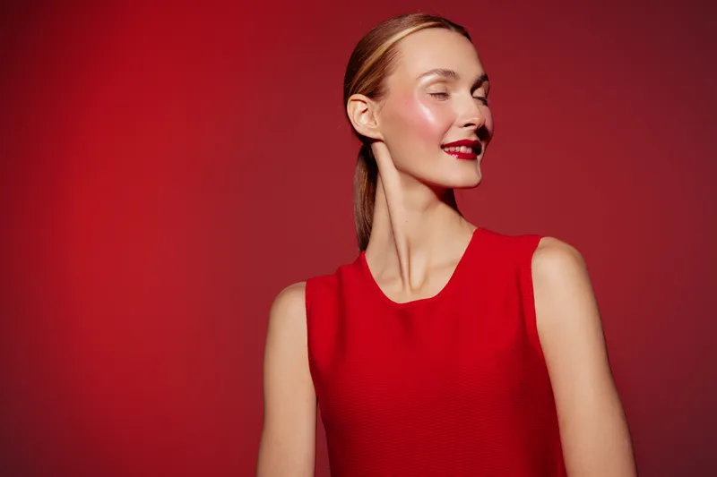 Smiling woman in red sleeveless dress poses against red background, looking sideways with closed eyes. Concept of fashion, beauty, confidence, and elegance in studio lighting.
