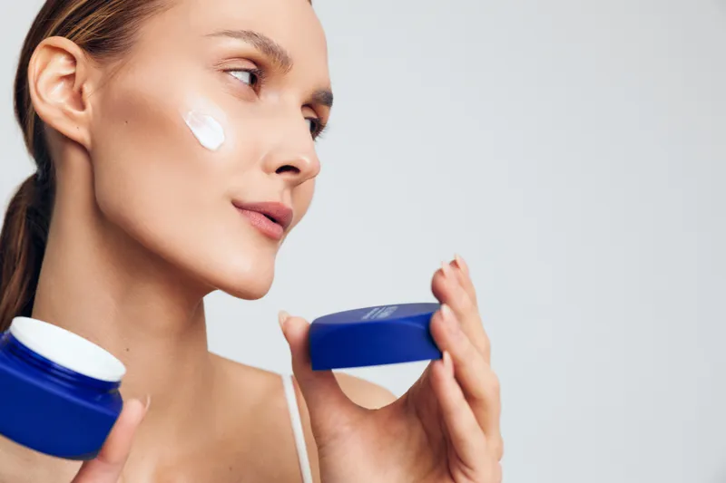 Young woman applying white face cream on cheek while holding blue cosmetic jar. Close-up portrait with soft lighting on light background. Concept of skincare, beauty, and self-care routine.