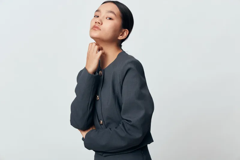Young woman with dark hair and serious expression posing in gray jacket against light background. Fashion, studio portrait and confident female model in minimalistic style for editorial use.