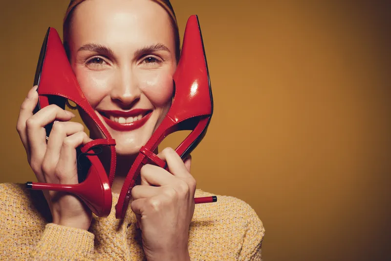 Smiling woman holds red high heels near face, wearing yellow sweater. Studio portrait with warm lighting on brown background. Concept of fashion, style, and happiness.