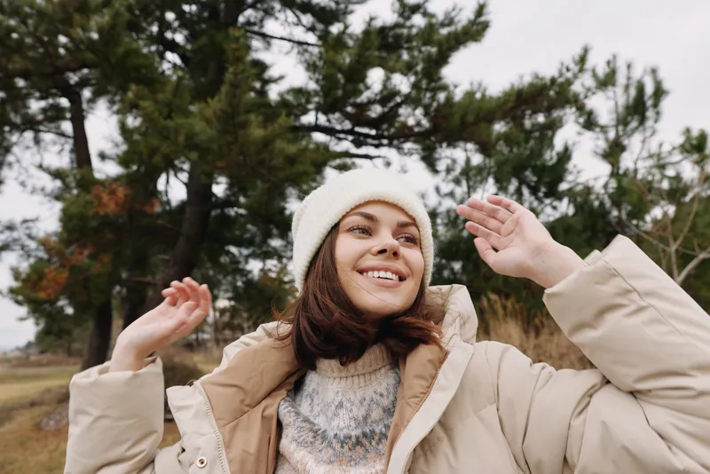 Happy woman outdoors wearing a beige jacket and warm winter coat smiles in a park setting, cozy attire and soft knit textures, neutral tones and relaxed pose, outdoor photography lifestyle shot
