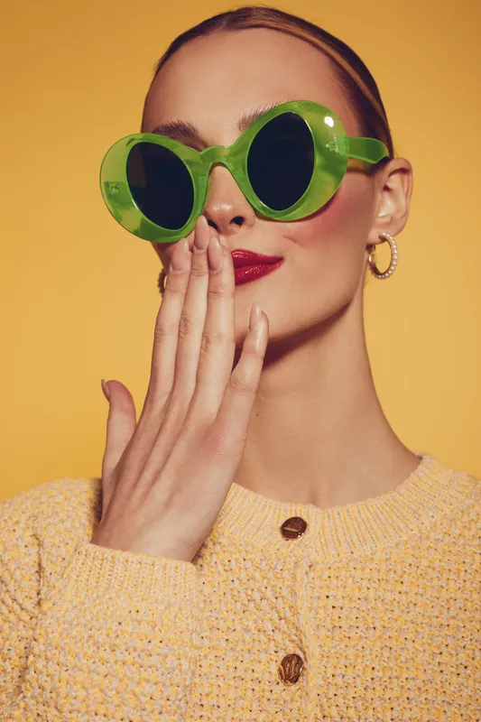 Stylish woman with green round sunglasses and red lipstick covers mouth with hand. She wears yellow knitted sweater and gold hoop earrings on yellow background. Fashion and beauty concept.