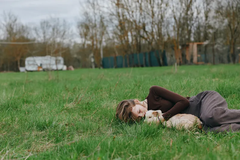 A relaxed woman lies beside a playful goat kid in a bright grassy field. A moment of rural lifestyle and gentle bonding outdoors, capturing calm, companionship, and simple joy in nature.