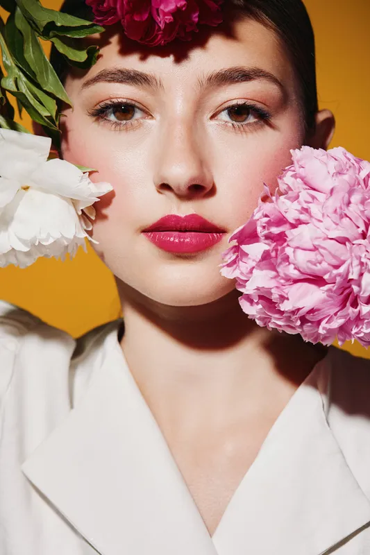 Young woman with fresh, glowing skin and vibrant pink lips poses holding colorful flowers near her face. Concept of beauty, fashion, and creative floral styling on plain background.