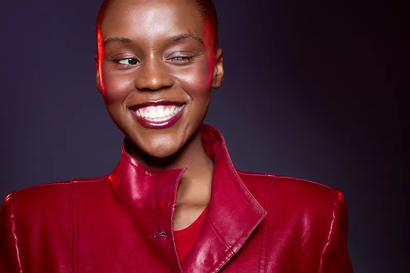 Smiling African woman with dark skin wearing shiny red leather jacket and lipstick looks aside against dark background. Concept of beauty, fashion, confidence, and style for black models.