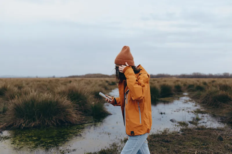 Outdoors woman lifestyle, wearing coat, and scarf, walks a trail through autumn grass under cloudy sky, holding smartphone. Wet path, travel and wellbeing. Field scenery with puddle.