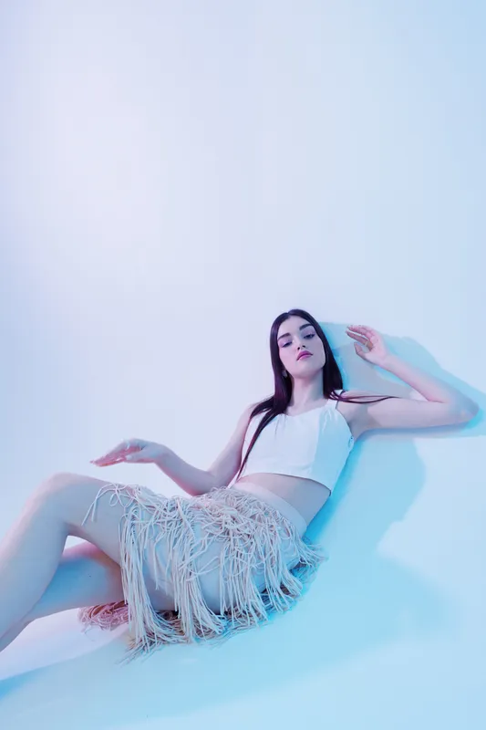 Young woman beauty fashion model poses lying down in a white top and fringed skirt on a light background. Concept for style, elegance, and modern clothing.