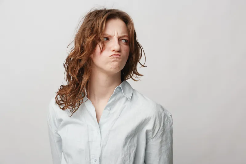 Young woman with curly hair wearing white shirt makes disgusted face looking away on plain background. Concept of dislike, displeasure, negative emotion, and facial expression.