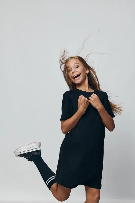 Girl jumping with joyful expression, wearing black dress and sneakers with striped socks, isolated on gray background with hair flying and playful mood in studio setting.