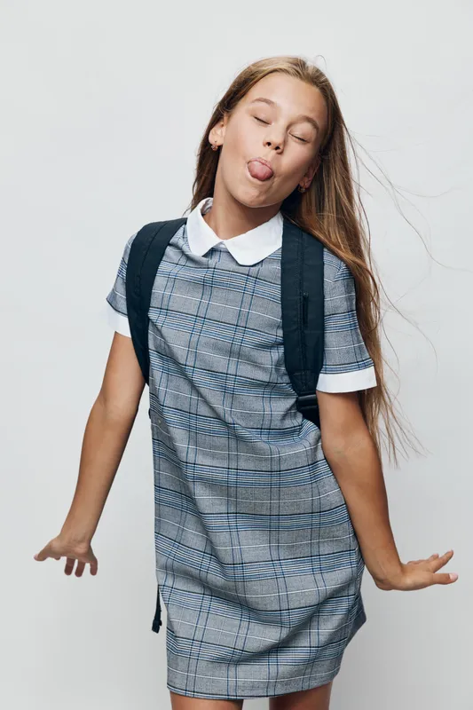 Girl with long hair wearing plaid dress and backpack making funny face sticking out tongue on white background. Child posing with closed eyes and playful gesture in studio.