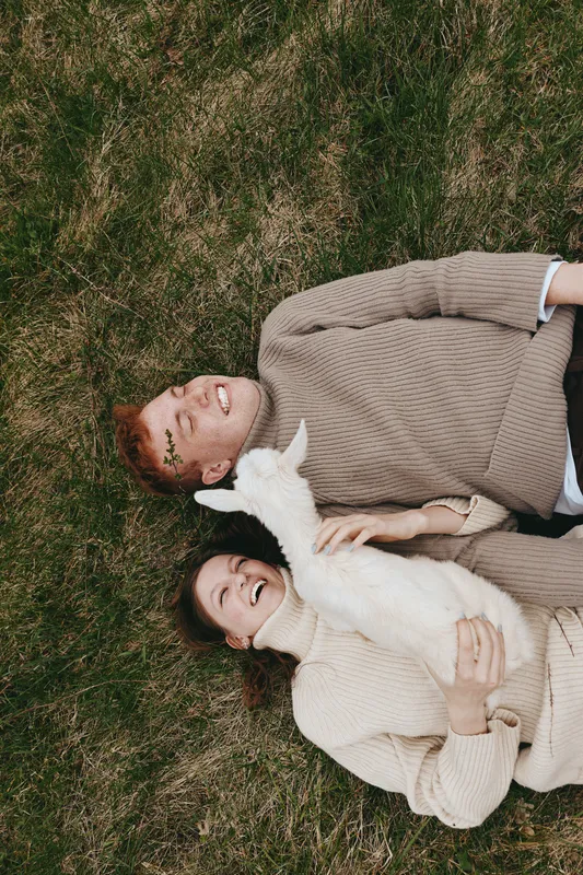 Young couple lying on grass smiling together while holding a baby goat in a cozy autumn setting with warm sweaters and natural light