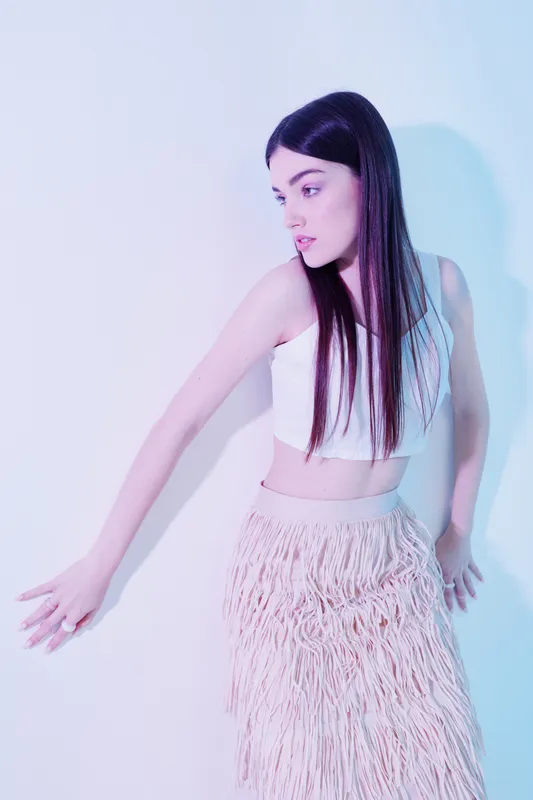 Young woman with long dark hair wearing a white crop top and textured skirt poses against a pastel background. Concept of beauty, fashion, and modern style.