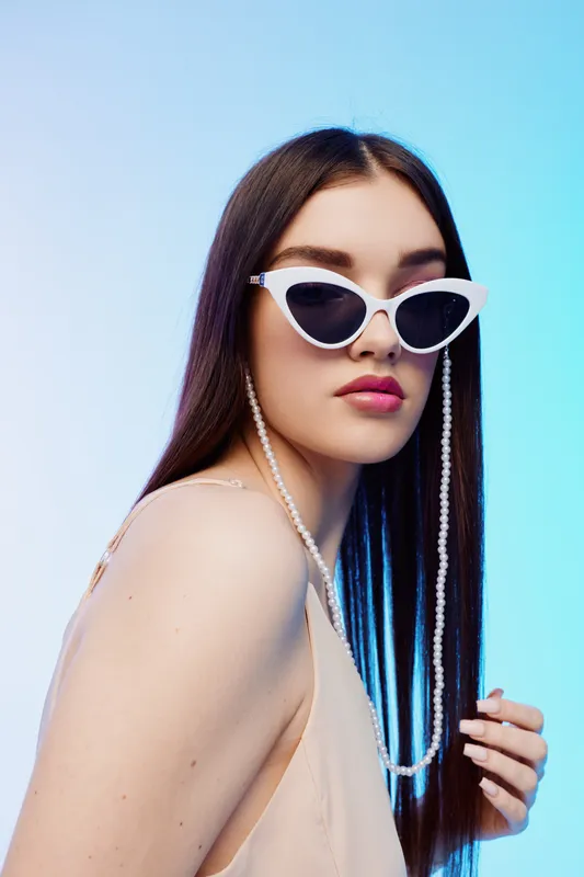 Young woman with long dark hair wears white sunglasses and a pearl chain. She poses against a gradient blue background. Concept of beauty, fashion, and style.