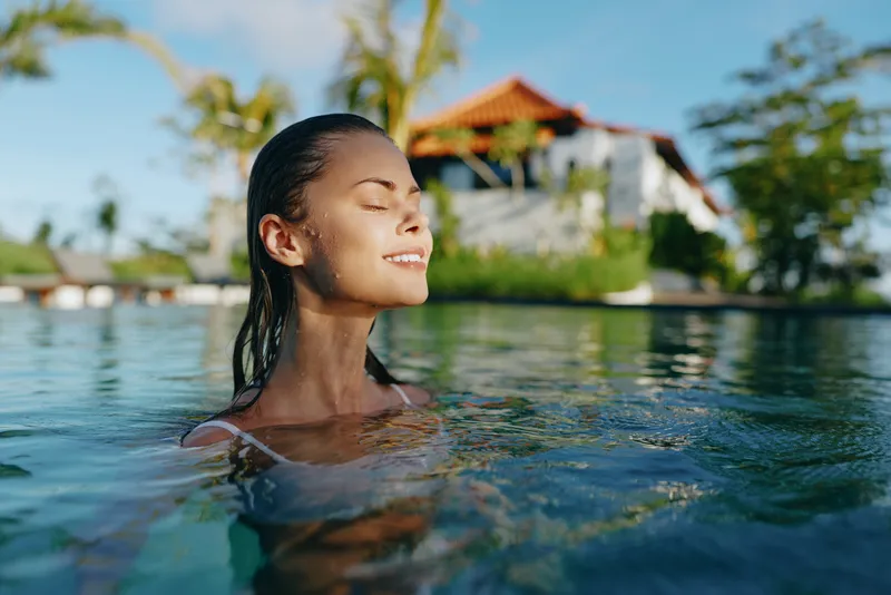 Cheerful woman relaxing in sunlit water, eyes closed, peaceful mood, tropical resort backdrop, clear reflections, gentle breeze, vibrant colors, carefree lifestyle