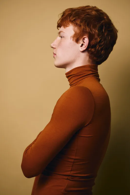 Young man with red hair wearing brown turtleneck sweater standing in profile with arms crossed against plain beige background. Calm expression with casual posture and natural lighting.