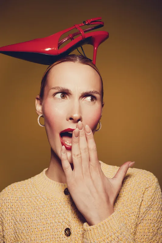 Surprised woman with red high heel shoe on head covers mouth with hand. Fashion concept with bold accessory and expressive facial expression on warm brown background.