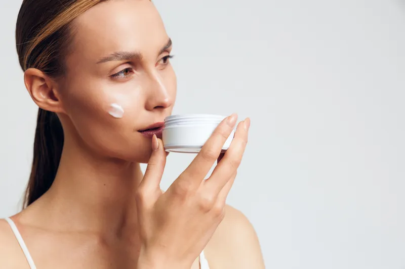 Young woman applying cream on her face, skincare routine in a bright studio, soft light, natural tones, calm moment, healthy glow, minimal makeup and serene ambience around soft vibe