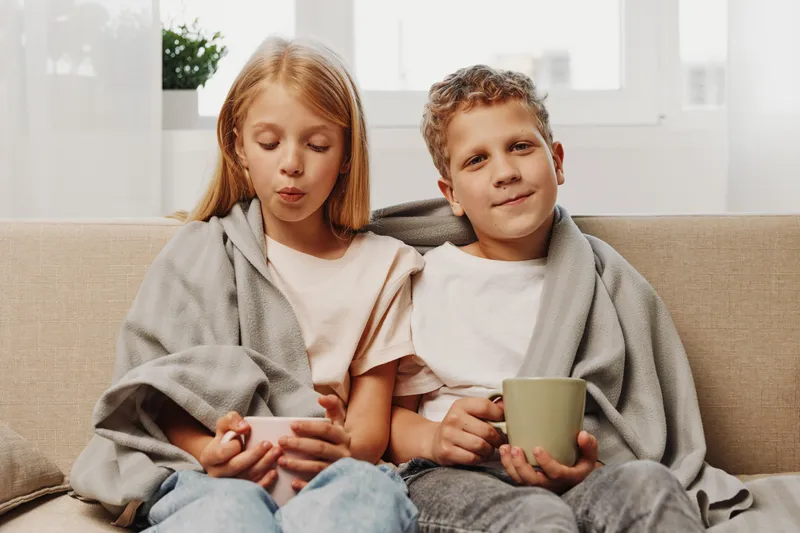 Two children wrapped in a grey blanket sit on a beige sofa holding white and green mugs, enjoying a cozy moment indoors by the bright window.