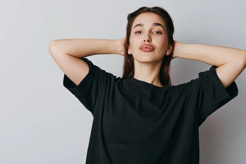 Young woman in black t shirt posing with hands behind head looking confident and relaxed against plain light background in casual style portrait photography.