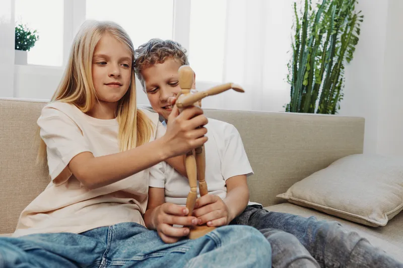 Two children sitting on a sofa play with a wooden artist mannequin together in a bright living room with modern plants in the background. Concept of creativity and childhood.