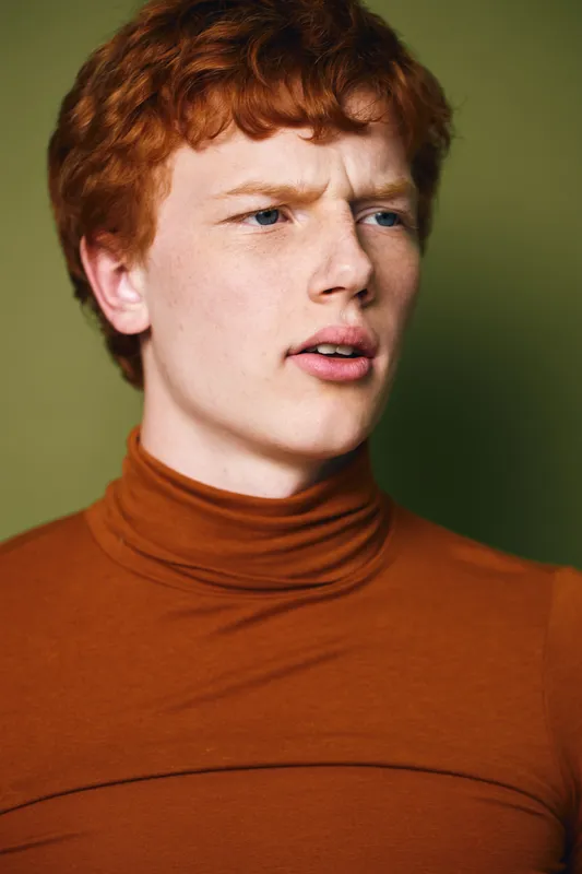 Young man with red hair and fair skin wearing brown turtleneck looks thoughtfully away with slightly open mouth against green background. Casual portrait with serious expression.