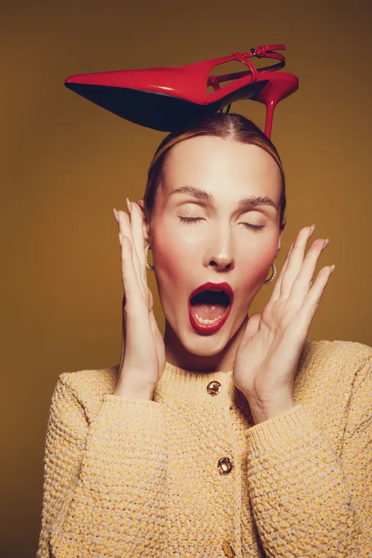 Young woman with closed eyes and open mouth holds hands near face while balancing red high heel shoe on head. Fashion concept with expressive emotion and stylish outfit on brown background.