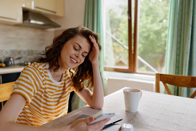 Cheerful woman at home, relaxed smile, striped shirt, seated at a table by a window, using a smartphone, warm natural light, cozy kitchen setting, casual lifestyle moment