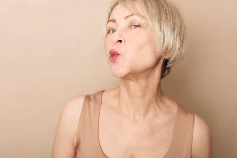 Blonde mature woman with short hair puckers lips making a kiss gesture on beige background. Concept of beauty, aging, confidence, and playful expression in natural light.