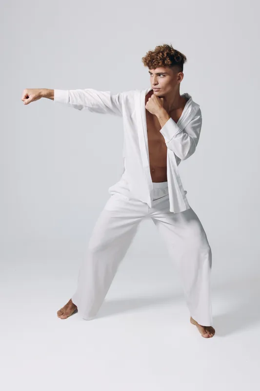 Young man practicing martial arts in white clothing with bare chest in studio. Athlete training punch stance with focused expression and balanced posture on plain background.