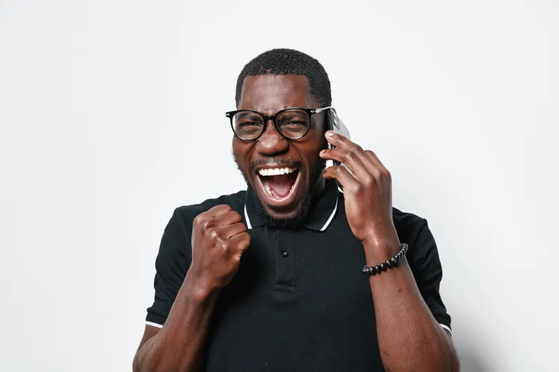 African American man with dark skin wearing glasses and black polo shirt talking on phone and celebrating success with fist pump on white background.