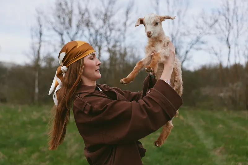 Young woman with long hair and headscarf holds a baby goat in a green field during an overcast day, capturing a tender moment of connection and lifestyle outdoors.