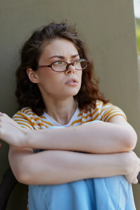 Pensive woman with glasses sits against a wall outdoors, arms crossed on her knees, wearing a striped shirt and jeans, taking a quiet contemplative moment.