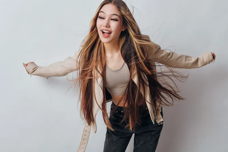 Young woman with long hair and casual outfit posing with arms outstretched on grey background. Female model smiling and expressing freedom, movement and happiness indoors.
